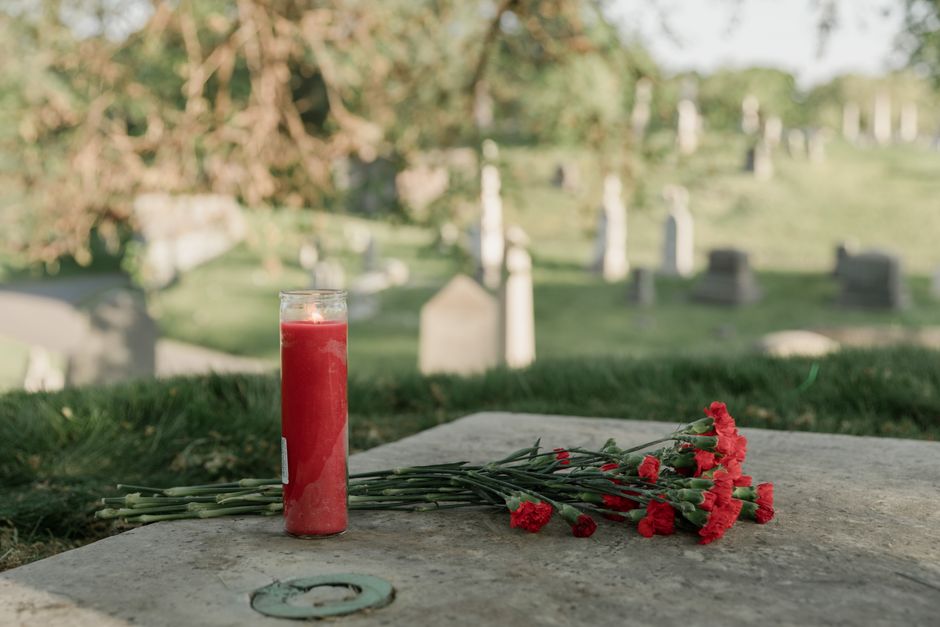 A tall glass candle holder filled with red wax, placed on a stone surface, beside a small bouquet of red carnations with green stems and buds, lying on the same stone. The scene is set outdoors in a c