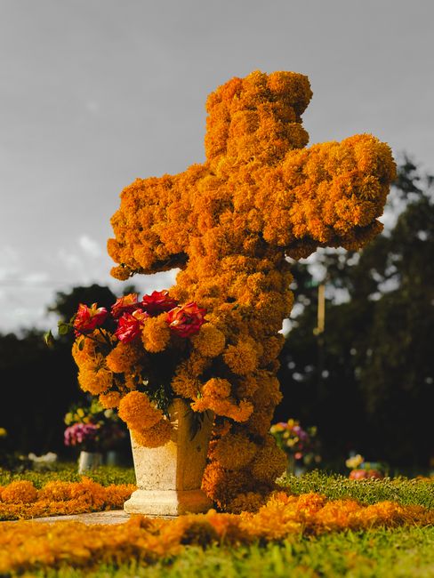 A floral cross monument made entirely of densely packed bright orange marigold flowers, standing on a stone base in an outdoor setting. The arrangement is vertically oriented with the flowers forming 