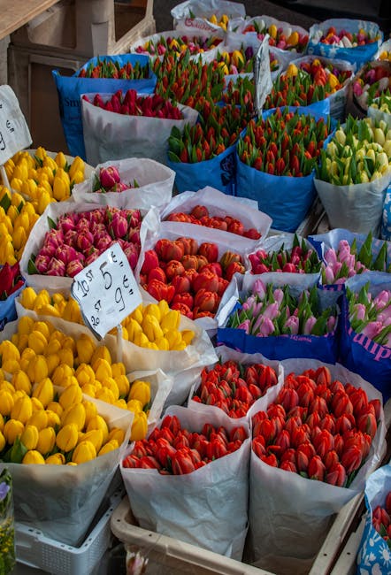 A vibrant display of freshly cut tulips arranged in multiple large paper-wrapped bouquets at a flower market. The tulips are in various bright colors, including yellow, pink, red, and green, with some