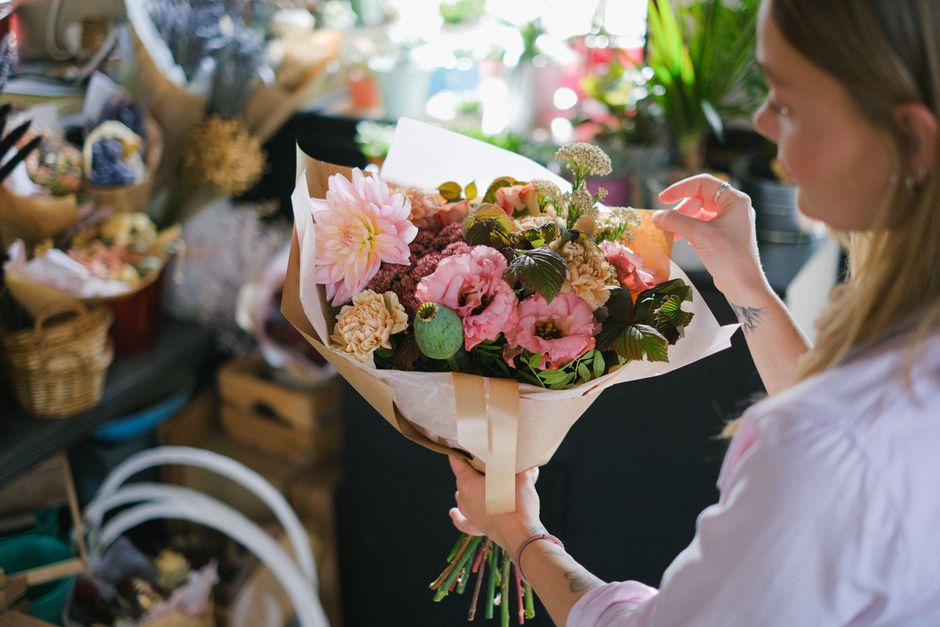 A woman with long, light brown hair holding a floral bouquet featuring large pink dahlias, soft pink lisianthus, cream-colored carnations, and greenery wrapped in light brown paper with a beige ribbon