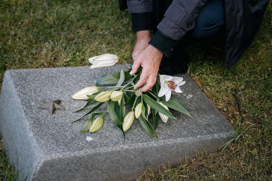 A person placing a floral arrangement of white and cream lilies with elongated green leaves on a polished grey granite memorial stone. The lilies include several fully closed buds and one open bloom d
