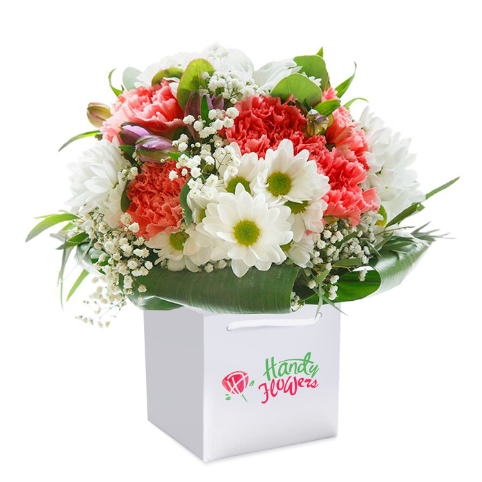 Captured in soft, even lighting, a luxurious bouquet rests perfectly upright in a minimalist square white gift bag, poised for delivery across North Ockendon. Ruffled coral and deep red carnations form the heart of the arrangement, their petals abundant and artfully curled, conveying a feeling of richness and affection. Nestled among them are clusters of bright white daisies, their lime-yellow centers glowing with a sunlit optimism, just like the community spirit you meet near Peartree Green. Featherlight sprays of white baby's breath are woven throughout, infusing the bouquet with an ethereal airiness that contrasts with the lush petals and grounding foliage. Emerging softly from the left are small, closed purple buds, perhaps alstroemeria, adding a hint of intrigue and depth. Cascading around the base, broad and glossy green leaves form a natural collar, highlighting the meticulous handwork that local North Ockendon florists put into every design. The white gift bag's pristine finish, complete with sturdy ribbon handles, ensures the bouquet is as practical for transport as it is eye-catching-a sophisticated gesture, perfect for surprising loved ones at home or work. Every element of this image-from its color to its composition-radiates freshness, care, and the authentic artistry cherished throughout North Ockendon, reflecting the thoughtful touch delivered with every order from Flower Delivery North Ockendon.
